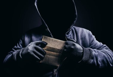 Photo of scary shaded hooded man hiding cardboard box parcel on dark background. clipart