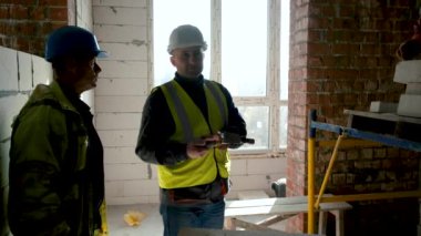 İnşaat alanındaki ilk güvenliği açıklayan işçi. İki işçi - sanayi mühendisleri kasket takıyor, depoda güvenlik önlüğü var.