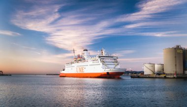 Günbatımında limana demir atmış büyük turuncu ve beyaz feribot. Arka planda endüstriyel yapılar görülebiliyor.