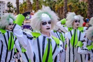 Çizgili siyah beyaz kostümler giymiş neon yeşili aksanlı bir grup insan. Beyaz peruklar takıyorlar ve yüzleri beyaza boyanmış. Gözlerinin etrafında koyu aksanlar var.