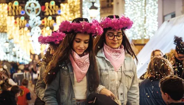 stock image two girl walking through what appears to be an outdoor festive environment, such as a Christmas market or similar holiday event. They are wearing matching pink feathered headbands and pink scarves