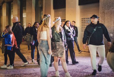 city street at night. Some people are standing and appear to be talking amongst themselves, while others are walking, likely passing by. The environment an urban setting with buildings lining the clipart