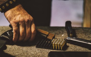 The image shows a person handling ammunition. There is a hand holding a handgun magazine, with a tray of bullets and parts of a firearm visible on a surface. The environment appears to be related to clipart