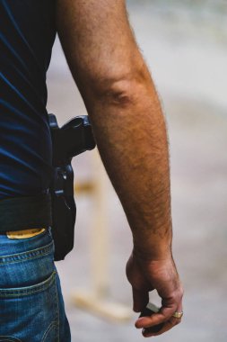 Görüntü, bir kişinin kol ve kalça bölgesinin yakın çekimini gösteriyor. Kotlu ve kemerine tabanca bağlanmış bir kılıfı var. El bir nesne tutuyor gibi görünüyor ama öyle değil.