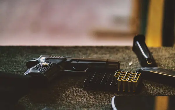 Resimde bir tabanca, birkaç mermi ve bir şarjör yer alıyor. Mekan kapalı bir ortam gibi görünüyor, muhtemelen bir atış poligonu ya da ateşli silahların kullanıldığı bir alan.