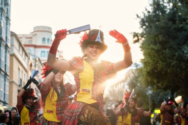 The image depicts a lively street scene with a group of people dressed in colorful costumes. The central figure is a smiling person wearing a hat resembling a top hat with a decorative addition that clipart