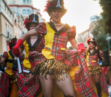 The image shows a group of people dressed in colorful, coordinated costumes, featuring a plaid tartan pattern. They are wearing hats, have bright yellow and red tops with frilly details, and layered clipart