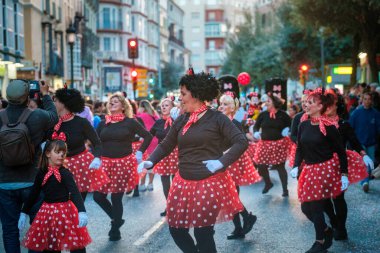 The image shows a group of people participating in what appears to be a street parade or festival. They are dressed in matching costumes featuring red skirts with white polka dots, black tops, and red clipart