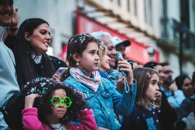 Bu resim bir grup insanı gösteriyor, muhtemelen halka açık bir etkinlikte ya da toplantıda. Çocuklar ve yetişkinler ortada, bir kız akıllı telefonu tutuyor, muhtemelen kaydediyor ya da alıyor.