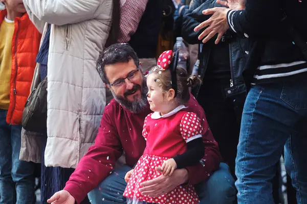 Görüntü bir adam ve genç bir kızı tasvir ediyor. Adam oturuyor, gözlükleri ve sakalı var. Kırmızı gömlek ve mavi kot pantolon giyiyor. Genç kıza nazik bir ifadeyle bakıyor, muhtemelen.