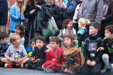 Çocuklar aile dostu bir açık hava etkinliğinde yetişkinler tarafından çevrelenmiş kostümlü bir gösteriyi hevesle izliyorlar..
