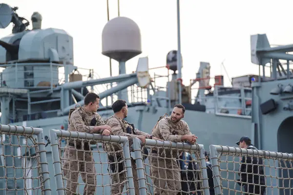 İspanya Malaga 128 2024 Askeri personel grubu şafak vakti bir donanma gemisinde kamuflaj üniformaları içinde, tartışmalı, güvenlik ağı ile korunuyor.