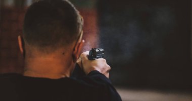 Görüntü, arkadan tabancaya nişan alan birini gösteriyor. Odak noktası, ellerinde görünen ateşli silah ve ortam loş bir ışık gibi görünüyor. İçerik bir vurulma olduğunu gösteriyor.