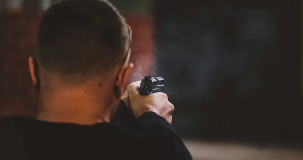 Görüntü, arkadan tabancaya nişan alan birini gösteriyor. Odak noktası, ellerinde görünen ateşli silah ve ortam loş bir ışık gibi görünüyor. İçerik bir vurulma olduğunu gösteriyor.