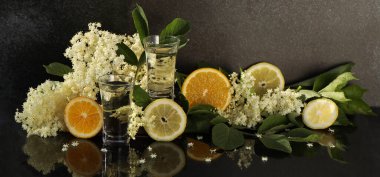 Yaşlı çiçeklerin süslendiği ve siyah arka planda dilimlenmiş turunçgiller olan mürver likörü.