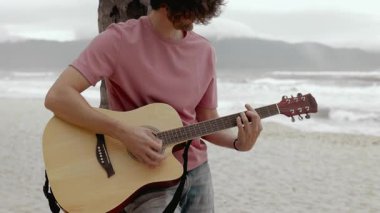 Sahilde gitar çalan genç müzisyen okyanus dalgaları ve manzaralı dağlarla