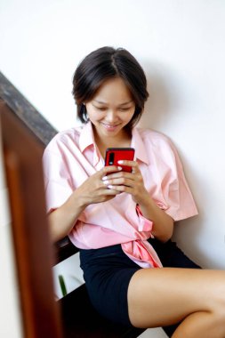 Kapalı alanda akıllı telefondan mesaj atarken gülümseyen Asyalı genç bayan..