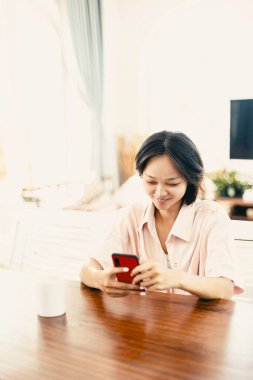 Parlak oturma odasındaki ahşap masada oturan Asyalı genç kadın telefona gülümsüyor..