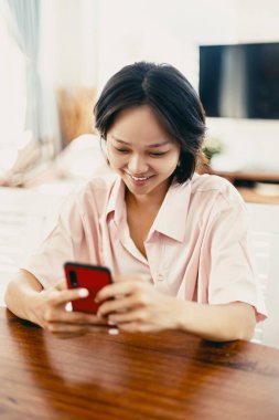 Genç Asyalı kadın evde akıllı telefon kullanırken gülümsüyor..