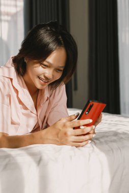 Yatakta kırmızı akıllı telefon kullanırken gülümseyen Asyalı genç kadın..