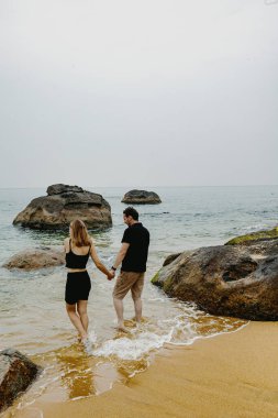 Genç beyaz bir çift kayalık bir sahilde el ele yürüyor..