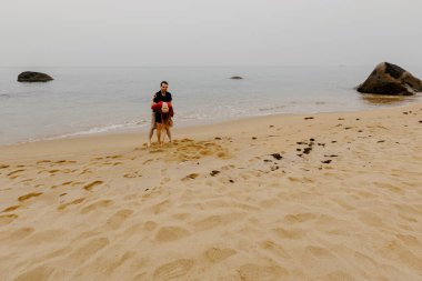 Genç beyaz erkek, arka planda köpek ve okyanusla kumsalda gezinti yapıyor..