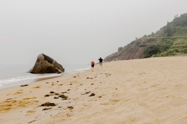 Arka planda kaya ve kayalıklarla sisli bir sahil yürüyüşü yapan çift..