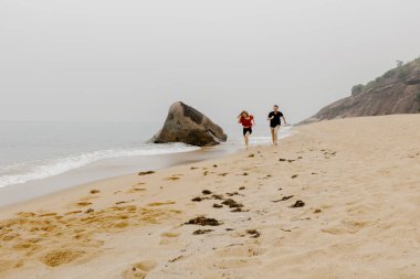Okyanus dalgaları ve kayalarla kumsalda koşan iki kişi..