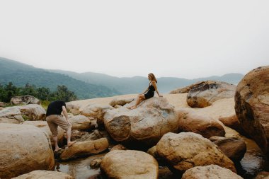 Genç beyaz kadın dağlık arazide kayaların üzerinde dinleniyor, erkek dereyi keşfediyor..