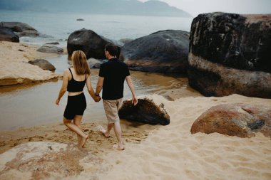 Gün batımında kayalık bir plajda el ele yürüyen beyaz bir çift..