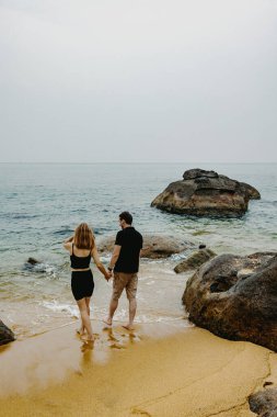 Genç beyaz bir çift kayalık bir sahilde el ele yürüyor..