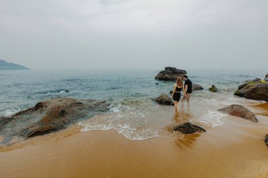Genç beyaz çift bulutlu gökyüzünün altında kayalık sahil şeridinde yürüyor..