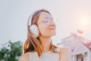 Genç beyaz kadın dışarıda müzik dinliyor kulaklıkları ve güneş gözlükleriyle.