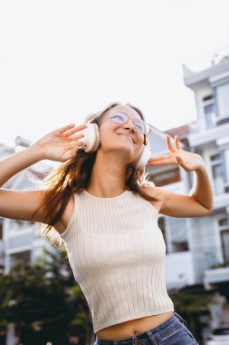Neşeli genç beyaz kadın kulaklıklarla dışarıda müzik dinliyor..
