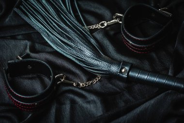 Siyah deri kamçı ve siyah kumaş arka planda kelepçeler..