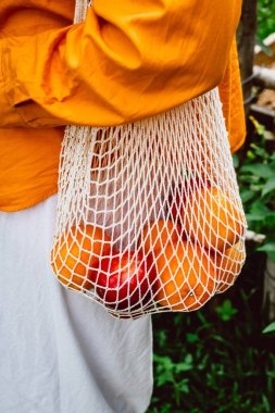 Taze portakallı ve kırmızı elmalı çevre dostu ağ torbası taşıyan kişi..