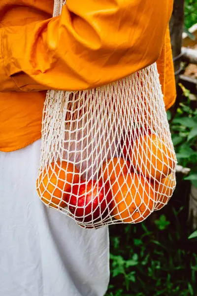 Taze portakallı ve kırmızı elmalı çevre dostu ağ torbası taşıyan kişi..