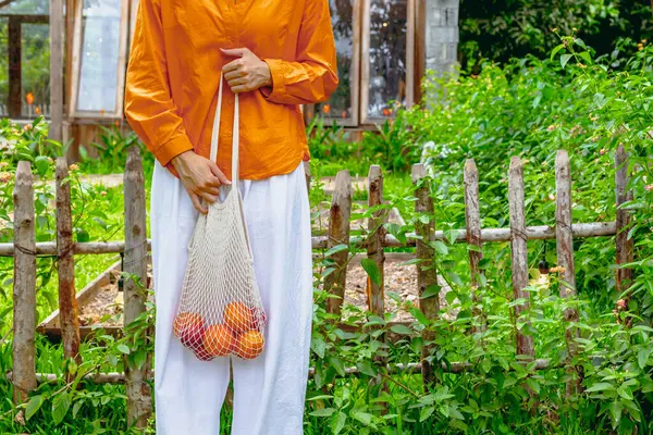 Bahçede taze ürünlerle dolu ağ torbası tutan Asyalı genç bir kadın..