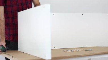 Siyah pantolon ve kırmızı kareli gömlek giymiş bir marangoz, beyaz bir kitaplık monte ediyor. 4K UHD