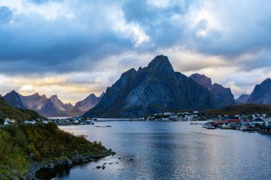 Norveç 'in kuzeyindeki Reine, Lofoten Adaları' nda sonbahar günbatımı, dramatik dağlar, sakin sular ve kıyı boyunca geleneksel evler..