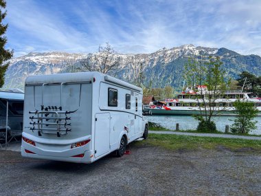 Beyaz karavan, arka planında feribot ve muhteşem İsviçre alpleri olan huzurlu bir göl kenarı kamp yerinin keyfini çıkarıyor.
