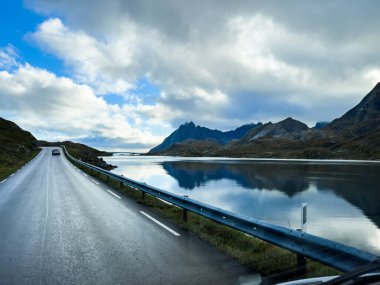 Norveç 'te bir fiyortta manzaralı bir yolda giden beyaz bir karavan. Arka planda dağlar ve bulutlu bir gökyüzü var.