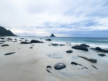 Norveç 'te bulutlu bir gökyüzünün altında beyaz kum ve kayaların turkuaz okyanus suyuyla buluştuğu güzel bir plaj manzarası.