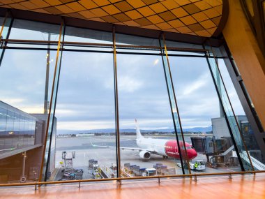Oslo, Norveç; 30 Eylül 2024: Kapıda park etmiş kırmızı ve beyaz bir uçak manzaralı geniş pencereli çağdaş havaalanı terminali
