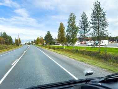 Rovaniemi, Finlandiya; 17 Eylül 2024: Güneşli bir sonbahar gününde, renkli ağaçlar ve mavi gökyüzü ile birlikte, beyaz çizgili asfalt bir yolun yanında, karavan park alanı