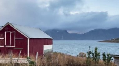 Rüzgâr Norveç fiyortlarında esiyor. Arkasında kırmızı ahşap bir ev ve dağlar var.