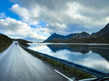 Norveç 'te bir fiyortta manzaralı bir yolda giden beyaz bir karavan. Arka planda dağlar ve bulutlu bir gökyüzü var.