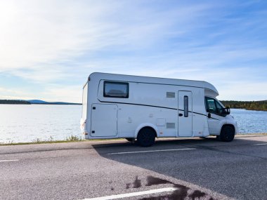 Güneşli bir günde gölün kenarına park edilmiş beyaz bir karavan manzarası. Yolculuk ve tatil için ideal.