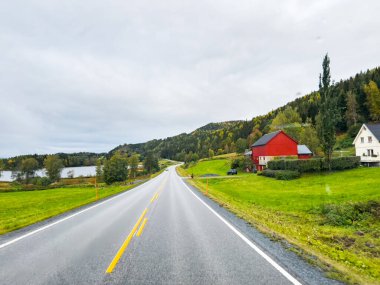 Norveç kırsalında: Geleneksel evleri ve huzurlu gölü olan, renkli bir sonbahar sahnesinde kıvrılan boş bir asfalt yol