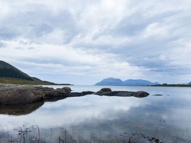 Sakin sular, sakin bir İskandinav fiyordunda bulutlu bir gökyüzünü yansıtıyor, heybetli dağlar, dingin atmosfere katıyor.